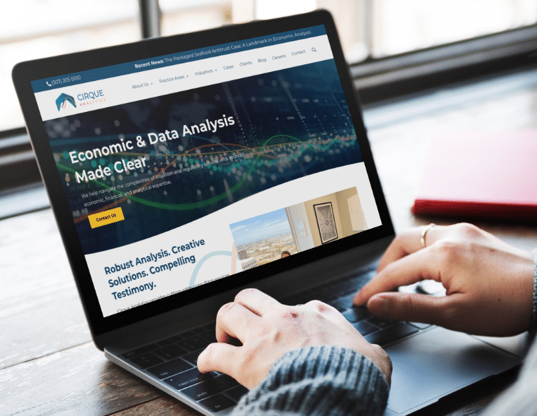 A person types on a laptop showing a website for Cirque Analytics, featuring economic and data analysis services. A red notebook and pen lie on the wooden table next to the laptop.